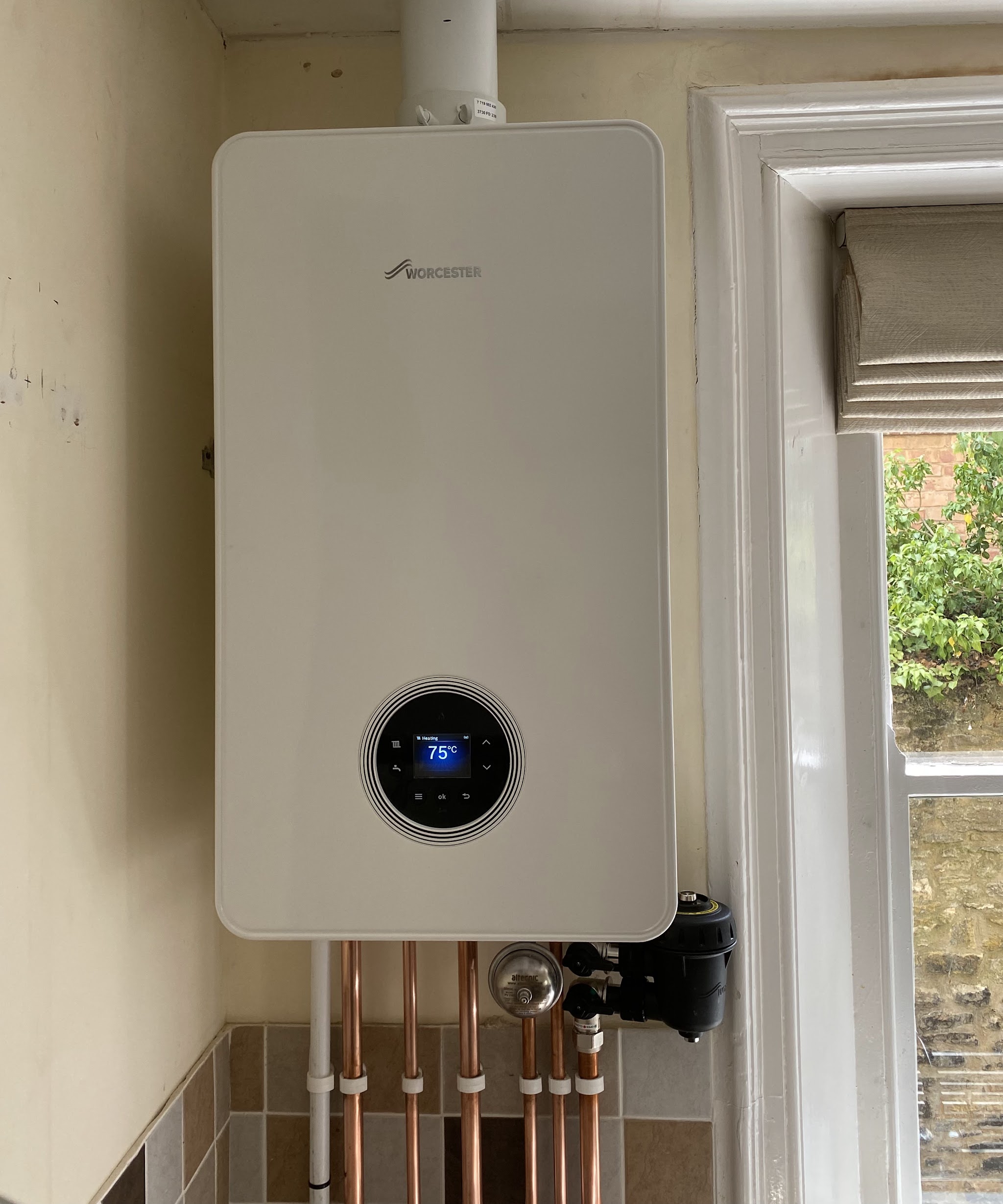 A new white combi boiler with a round display fitted beside a window, neat copper pipework below